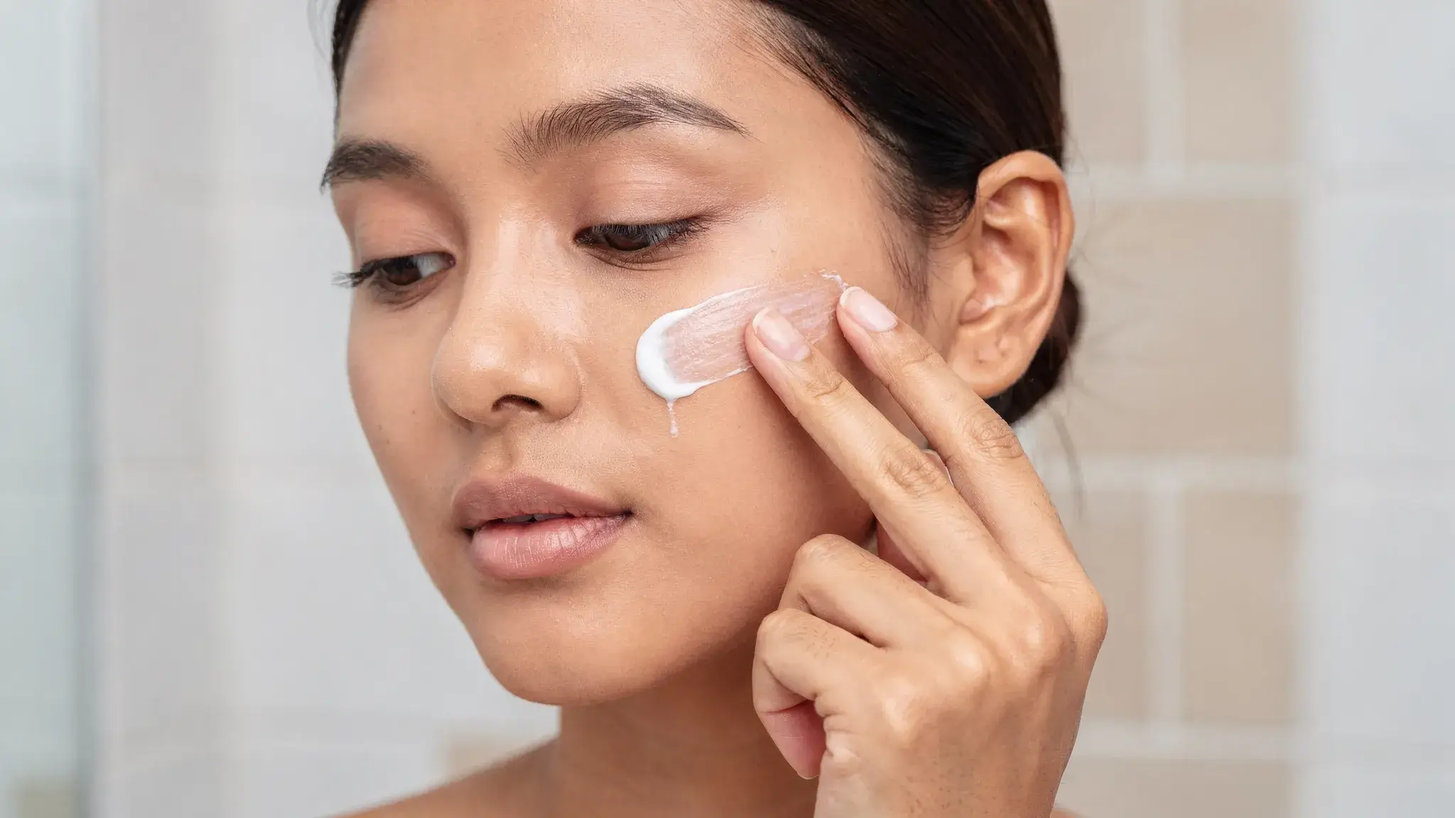 Close-up of a woman with medium skin tone applying a thin layer of depigmenting cream to her cheek in a bright bathroom, neutral lighting, no makeup, emphasizing careful fingertip application and a pea-sized amount.